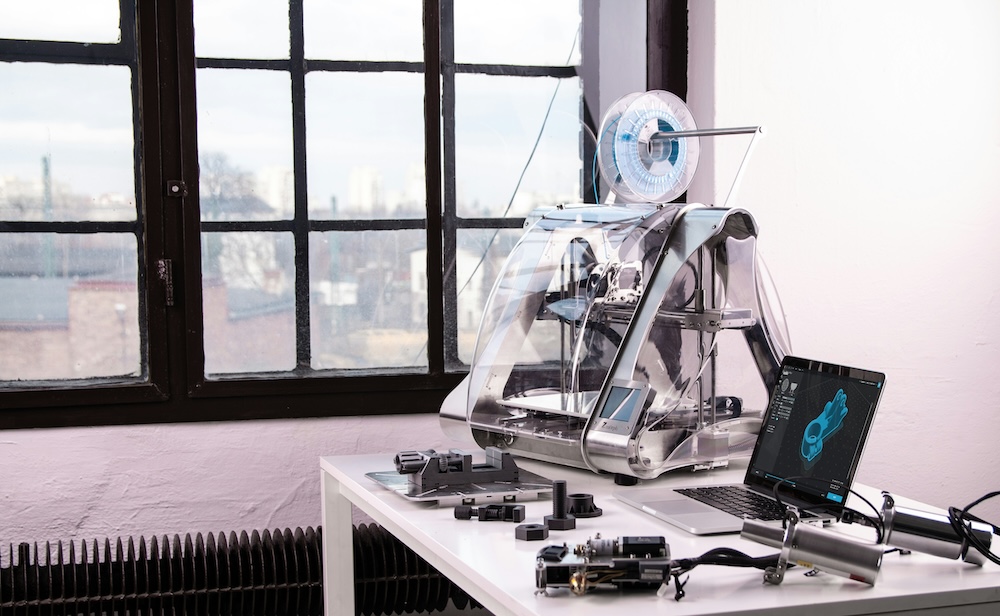 A 3D printer on a white desk with a laptop and tools.