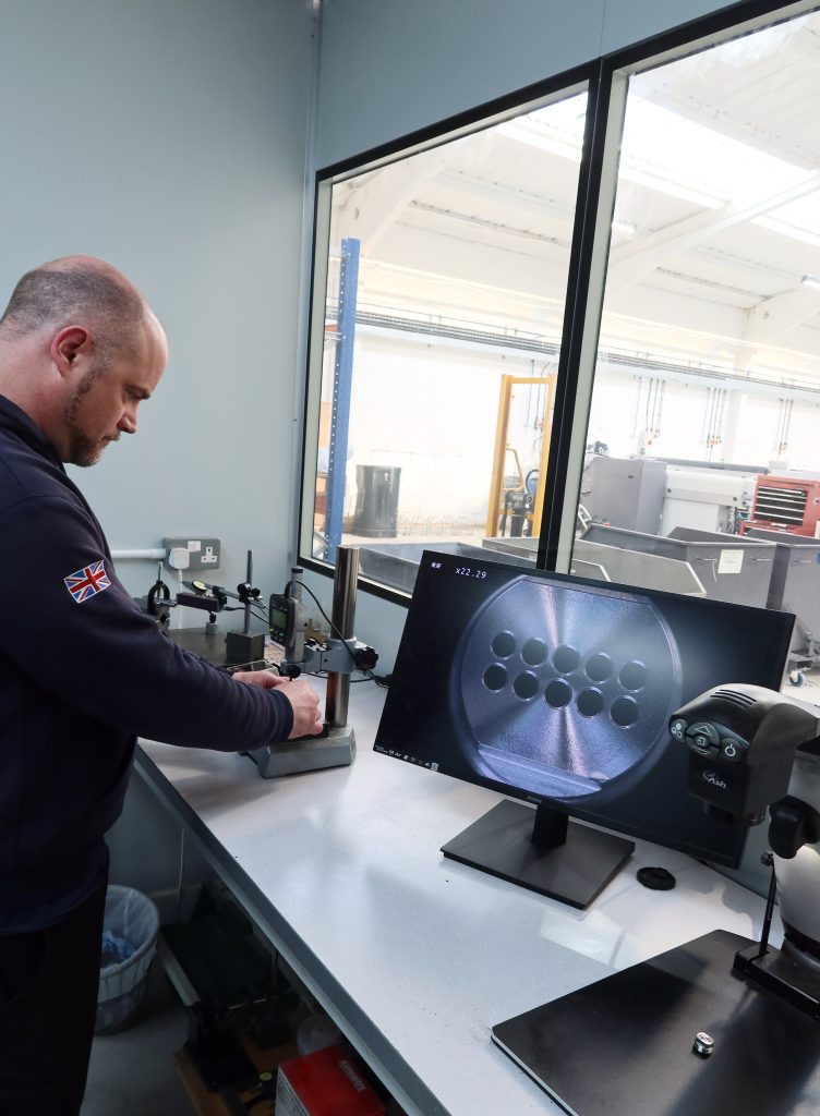 Sheldon Precision engineer using a digital vernier caliper to measure a precision-machined metal component in our workshop with CNC machinery in the background.